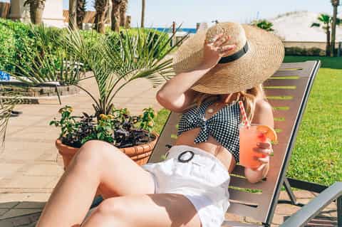 Woman relaxing on lounge chair wearing straw hat and checkered top, holding tropical drink in beachside setting