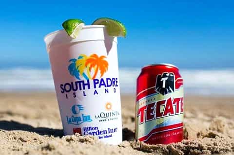 South Padre Island promotional cup and Tecate beer can on beach sand with ocean backdrop