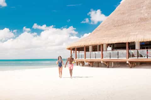 Couple walking on white sand beach toward overwater bungalow with thatched roof and turquoise ocean
