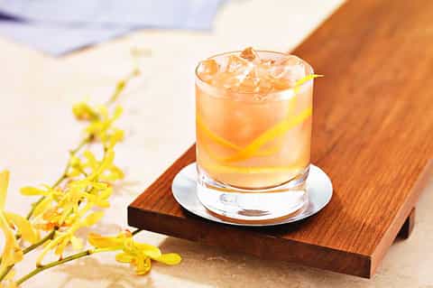 Amber cocktail with ice and lemon twist on wooden coaster with yellow flowers