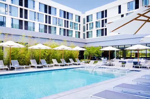 Modern hotel courtyard with rectangular pool, white loungers, umbrellas, and multi-story white building with uniform windows