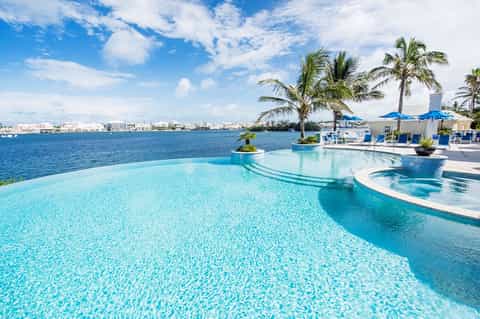 Luxury beachfront infinity pool overlooking ocean with palm trees, umbrellas, and coastal city skyline
