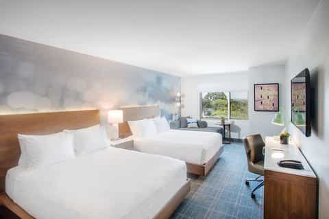 Modern hotel room with two queen beds, wooden headboards, work desk, and window with nature view