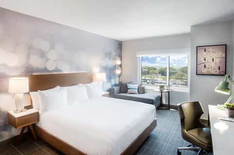 Contemporary hotel bedroom with queen bed, sitting area, and panoramic windows overlooking grounds
