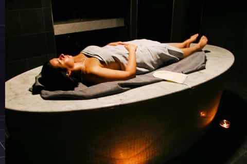 Woman relaxing in a dark stone spa bathtub with warm lighting and towels