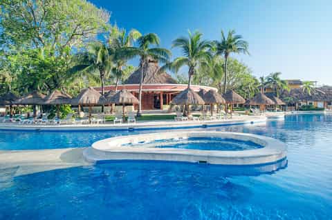 Resort lagoon pool with thatched-roof cabanas, lounge chairs, and palm trees under blue sky