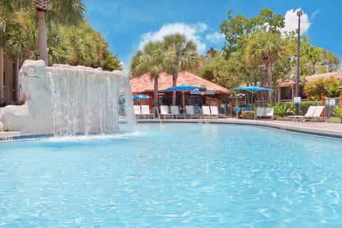 Resort pool with large stone waterfall feature, blue umbrellas, palm trees, and tropical landscaping