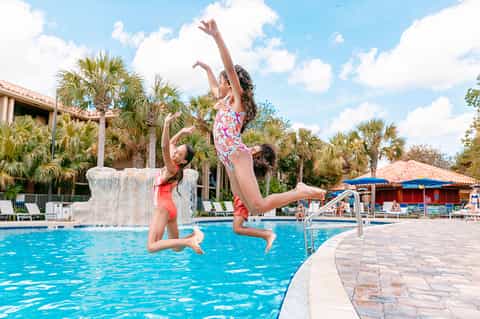 Two children jumping into a bright blue swimming pool with palm trees and cabanas in the background