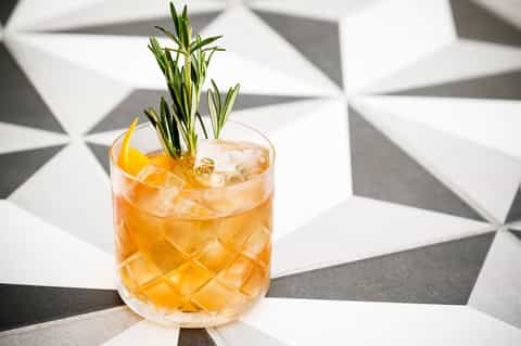 Golden whiskey cocktail with ice and fresh rosemary sprig on geometric patterned surface