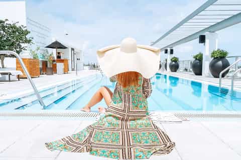 Woman in patterned dress and wide-brimmed hat relaxing poolside at a modern resort
