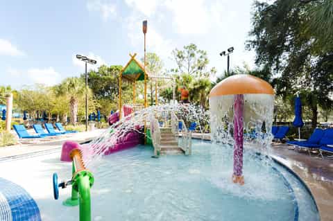 Colorful outdoor water park with splash pad, slides, and play structures surrounded by palm trees and lounge chairs