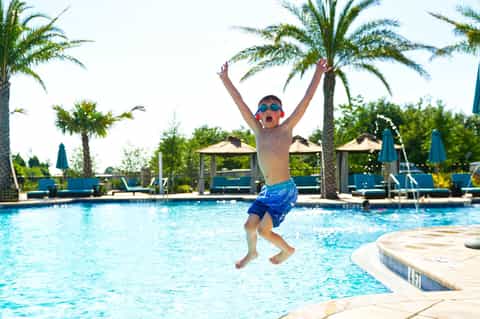 Child jumping into swimming pool with arms raised, surrounded by palm trees