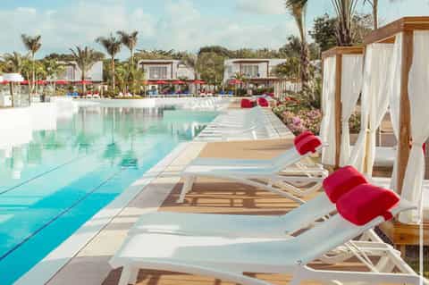 Resort pool deck with white loungers, wooden cabanas with white curtains, and palm trees