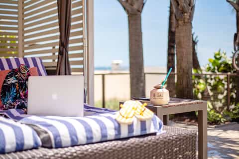 Laptop and tropical drink on striped lounger with palm trees and beachfront view