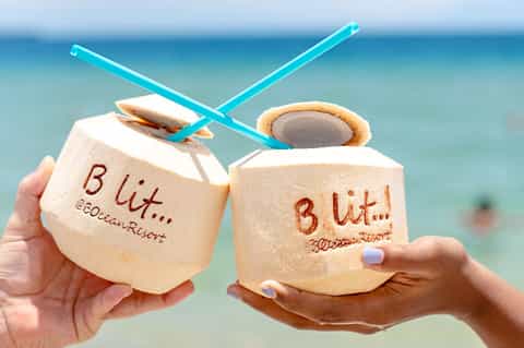 Two coconut drinks with blue straws clinking together against ocean backdrop at beach resort