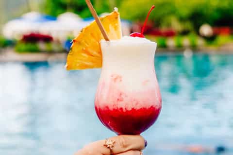 Tropical cocktail with red and white layered drink, pineapple slice, cherry, and straw by pool