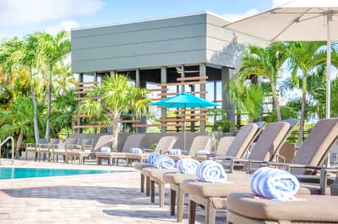 Luxury resort pool area with lounge chairs, blue umbrellas, palm trees, and modern architecture building