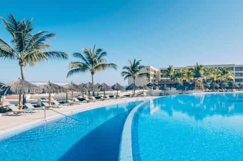 Large resort pool with palm trees, umbrellas, loungers, and beachfront hotel building