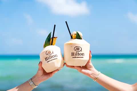 Hands holding two wooden coconut cocktails with Hilton branding against turquoise ocean