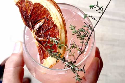 Grapefruit cocktail with pink liquid, fresh grapefruit slice, rosemary sprig, held in hand