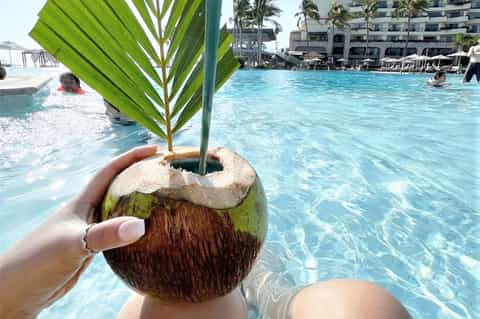 Person holding fresh coconut drink poolside with resort buildings and beach in background