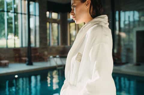 Woman in white spa robe standing by indoor swimming pool with modern architecture