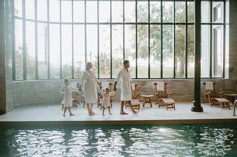 Family in white robes standing by indoor pool with large windows overlooking trees