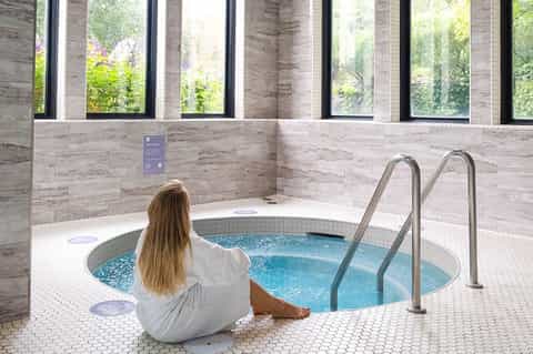 Woman in white robe sitting at edge of circular indoor hot tub with stone walls and large windows