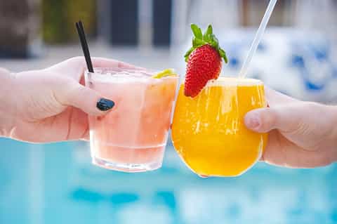 Two colorful tropical cocktails with fruit garnish held over turquoise pool water