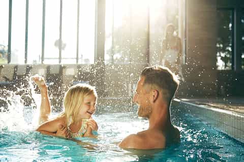 Indoor pool area with man and child swimming together, sunlight streaming through windows