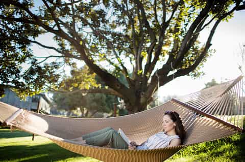 Woman relaxing in hammock under large tree with book in sunny backyard