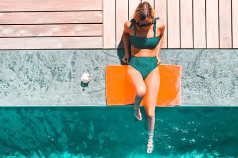 Woman in teal swimsuit sitting at pool edge about to enter turquoise water, aerial view