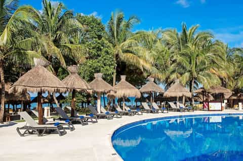 Beach resort pool area with thatched umbrellas, palm trees, lounge chairs, and ocean view