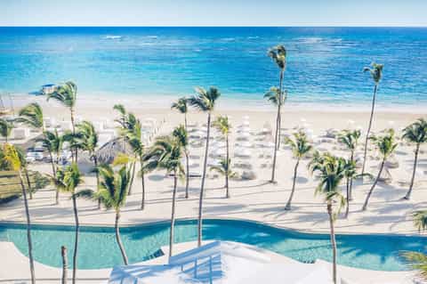 Aerial view of beach with palm trees, white umbrellas, pool, and turquoise ocean water
