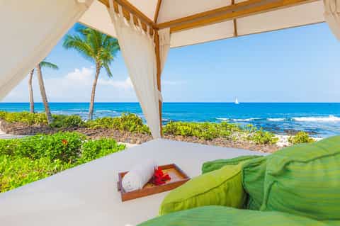 Beachfront cabana with ocean view, white curtains, palm trees, and sailboat on horizon
