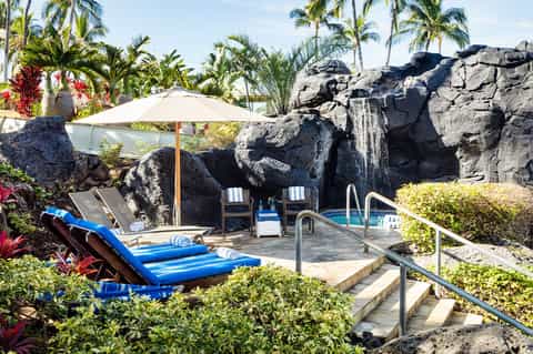 Resort pool area with blue loungers, white umbrella, and artificial rock waterfall features