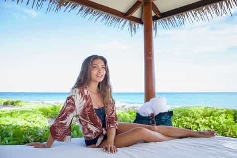 Woman in patterned jacket relaxing on beach platform under thatched umbrella with ocean view