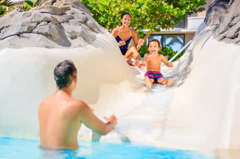 Family playing in shallow resort pool with large white boulder water slide feature