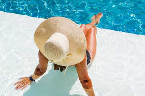 Person relaxing by turquoise pool wearing straw hat, showing sunny resort vacation poolside experience.