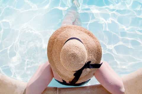 Person in pool wearing straw hat with black ribbon, viewing sunny water from poolside