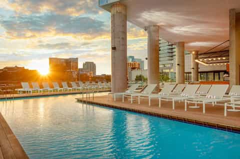 Indoor pool with natural light, white lounge chairs, and sunset city skyline views through large windows