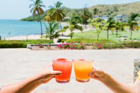 Two colorful drinks held up on beach with ocean, palm trees, and coastline in background