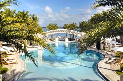 Luxurious resort pool with curved pergola, palm trees, loungers, and ocean view in background