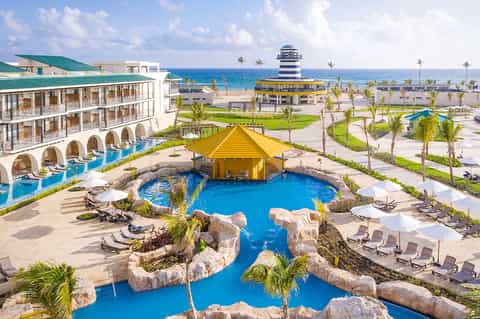 Beachfront resort with blue lazy river, yellow roof structure, palm trees, and ocean view