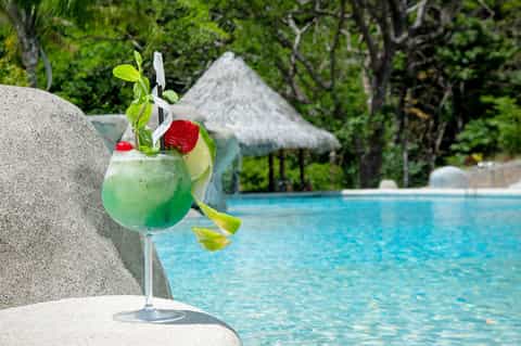 Green tropical cocktail with lime and mint garnish poolside with thatched roof cabana