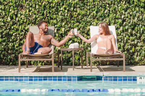 Couple relaxing in lounge chairs by the pool with green ivy wall backdrop, toasting drinks