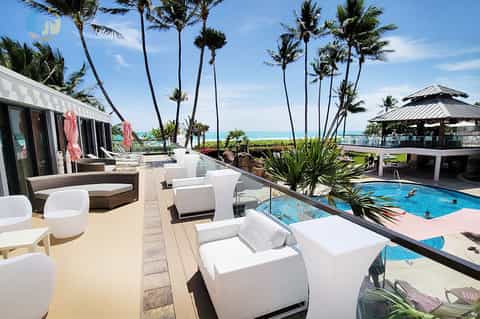 Beachfront resort pool deck with white lounge chairs, palm trees, ocean view, and modern architecture