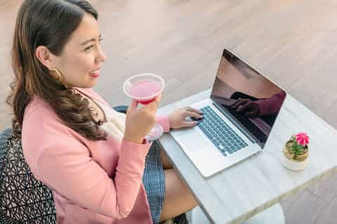 Woman working on a laptop while holding a pink cocktail at a modern desk with potted plant