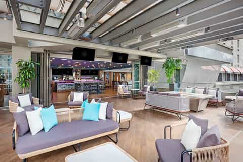 Modern resort lounge with purple seating, colorful pillows, wooden furniture, bar area, and indoor-outdoor layout with skylights