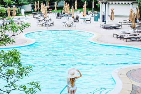 Resort pool area with crystal blue water, lounge chairs, tan umbrellas, and landscaped grounds with tropical greenery